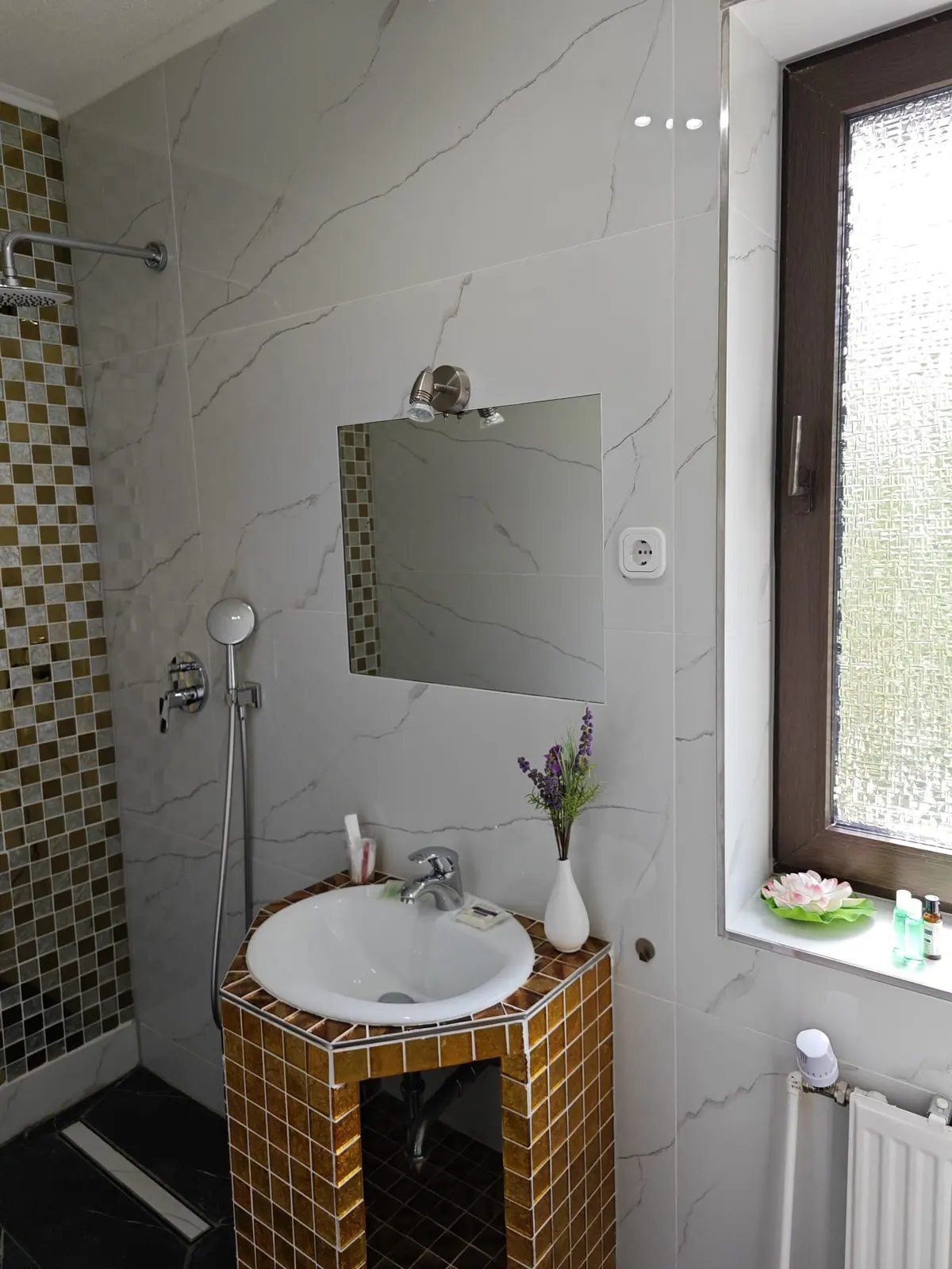Bad in Marmoroptik mit goldgerahmtem Spiegel, Waschbecken auf goldenem Mosaiksockel und Regendusche
