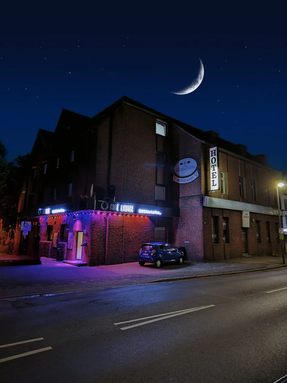 Jakz Hotel bei Nacht: Backsteinfassade an der Ewaldstraße mit dem Neonschriftzug Cafe Laurel und schmaler Mondsichel über dem Dach