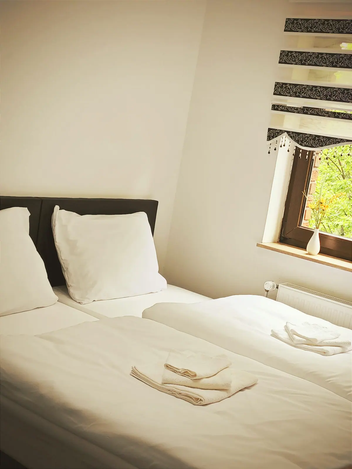 Zweibettzimmer mit Zebra-Rollo, Handtüchern auf beiden Betten und Blumen auf der Fensterbank